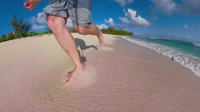 LOW ANGLE: Unrecognizable male tourist runs barefoot along the tropical beach in the sunny Caribbean. Excited man on vacation in Barbados runs along an empty white sand beach on a sunny summer day.