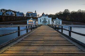 Fototapeta na wymiar Pier on the Baltic Sea. Illuminated wooden walkway on the island of Ruegen with a historic building in the evening for the autumn mood. Clear blue horizon over buildings in the background