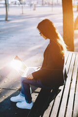 Young casual happy woman typing text on laptop while sitting on wooden bench, hipster girl using laptop outdoors, cross process image