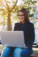 Portrait of young student girl in glasses sitting on the campus bench while use laptop gadget during her class break, attractive and elegant woman working on laptop sitting outdoors at sunny day