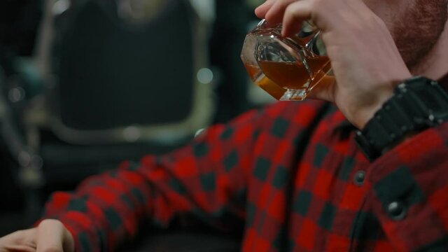A young guy waiting in line for a haircut takes a few sips of whiskey from the glass. Good customer attitude pepper service concept. Modern barbershop. 4k slow motion tracking shot.