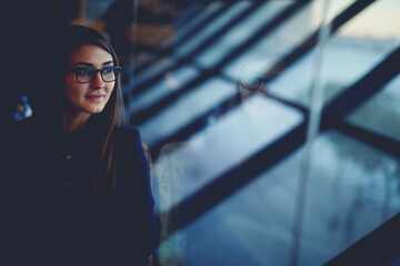 Portrait of charming woman in glasses looking out in big window while sitting in airport hall