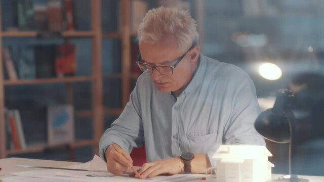 View through the glass window of concentrated senior architect drawing construction plan on paper with ruler and pencil and thinking about project while working at office desk in the evening