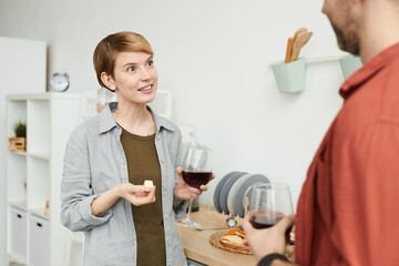 Young woman with short hair drinking red wine and eating cheese and talking to man while they standing in the kitchen