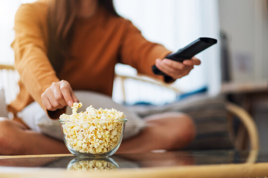 Close up image of a young woman eating popcorn and searching channel with remote control to watch tv while sitting on sofa at home