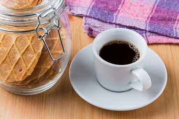 A cup of dark hot black coffee and a glass jar of waffle biscuits on a wood table