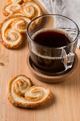 A glass cup of dark black hot coffee and some Palmier biscuits dropped around it on a wood table 