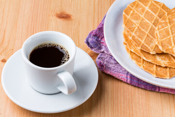 A cup os coffee and a pile of waffle biscuits in a plate on a wood table