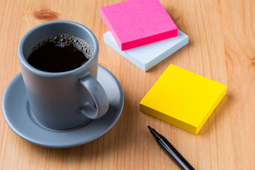 A light wood desktop table with a grey cup of coffee, a pen, and some colour note paper around
