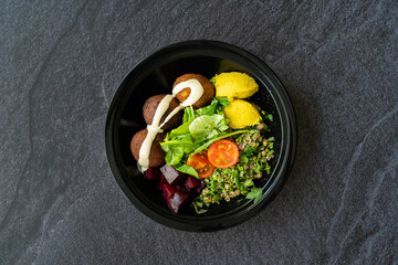 Healthy Vegan Take Away Falafel Bowl with Mung Bean Salad, Legume, Roasted Beet and Yellow Turmeric Hummus Tahini Sauce in Black Plastic Plate Cup Package / Container.