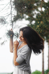 young attractive girl with long hair, brunette, in clothes of neutral colors, stands in the summer forest and holds a pine branch