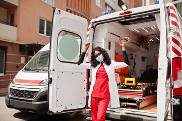 African american female paramedic in face protective medical mask standing in front of ambulance car.