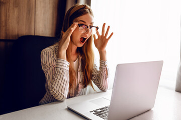 shocked, surprised young woman with glasses, working remotely on a laptop on the Internet, is sitting at home at a Desk. Quarantine, coronavirus