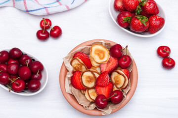 Mini tiny pancakes with straeberries and cherries on white wooden background. Trendy food concept. Flat lay.