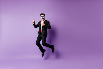Fototapeta na wymiar Excited well-dressed guy with scary makeup fooling around in studio. Zombie model funny jumping on purple background.
