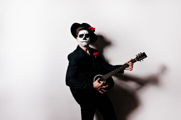 Studio portrait of mexican mariachi in deadman attire. Indoor photo of male model with zombie makeup playing guitar.