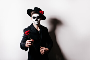 Fototapeta na wymiar Zombie man with rose posing in sombrero. Sad male model in black clothes standing on white background during halloween photoshoot.