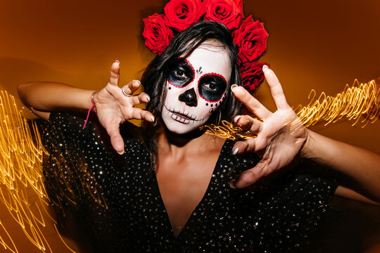 Gorgeous young female model with white face painting posing in halloween. Studio photo of debonair zombie girl standing on sparkle background.