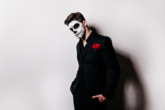 Handsome pensive man in black clothes waiting for halloween photoshoot. Indoor shot of enthusiastic zombie boy with face painting.
