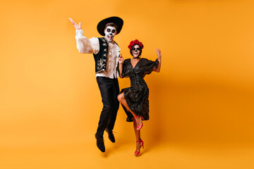 Fototapeta na wymiar Glad zombie man in sombrero jumping on yellow background. Charming muerte girl in black dress dancing with boyfriend.