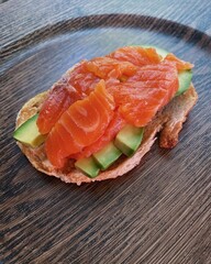 tasty fat aromatic sandwich with white fresh bread, croutons, avocado and salmon lies on a dark round wooden plate and stands on the table, top view