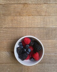 on a brown wooden planks there is a white bowl with different colored berries - blueberries, raspberries, blackberries, in soft light top view, flat lay