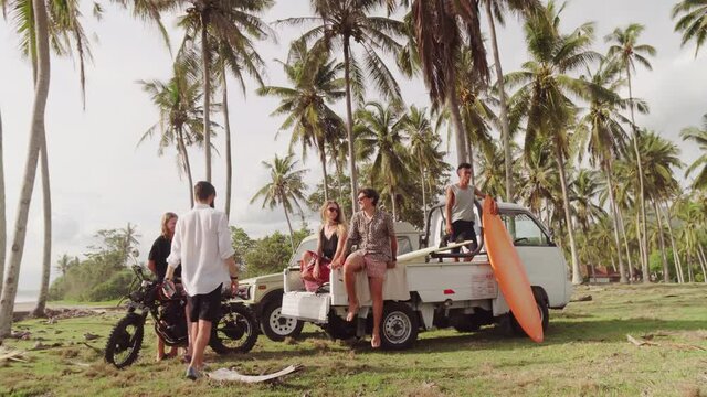 Wide shot with tracking of friends sitting on motorcycle and back of pickup truck while relaxing on coast in Bali
