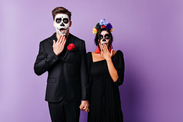 Shocked european couple with spooky makeup holding hands on purple background. Young people in black clothes posing in zombie attire in halloween.