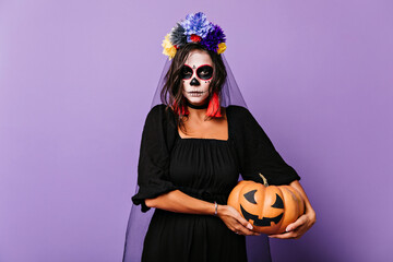 Serious brown-haired woman in veil holding halloween pumpkin. Indoor shot of gorgeous girl in dead...