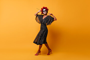 Full-length portrait of stunning female zombie in mexican attire. Inspired slim girl in halloween costume dancing on orange background.