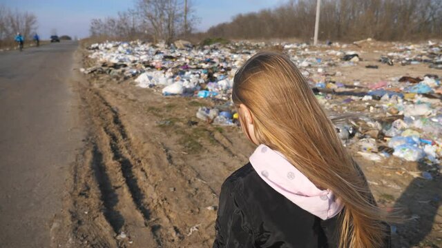 Little girl goes on the road against the background of garbage dump. Small female child with blonde hair walks along driveway near a lot of trash at countryside. Environmental pollution problem