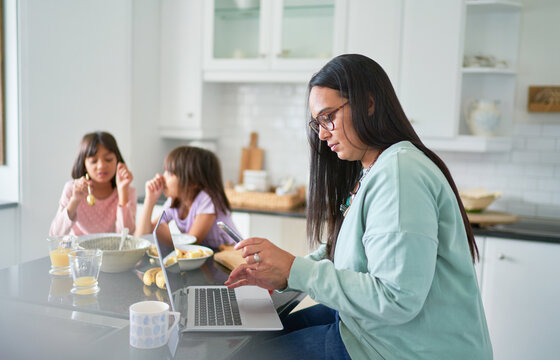 Mother working at laptop in kitchen while daughters eat breakfast