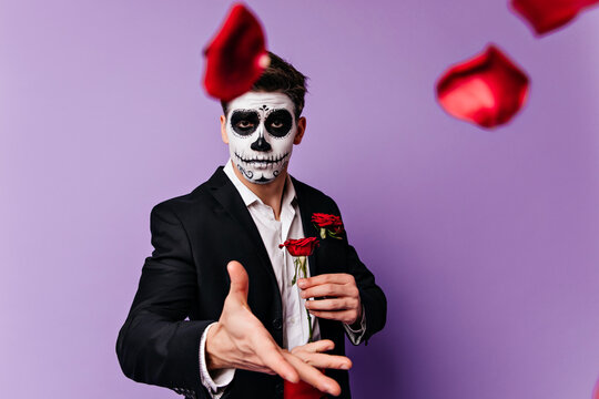 Sad european guy with spooky makeup throwing out rose petals. Indoor photo of dead man posing with flowers in halloween.