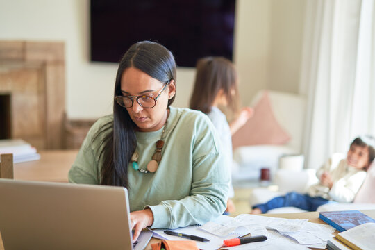 Mother paying bills on laptop with kids playing in background