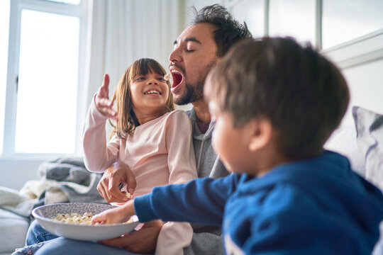 Playful kids feeding popcorn to their father on sofa at home