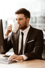 Business concept. Successful young businessman at work. Manager sitting at the office table happy drinking coffee from cup. Man smiling in suit indoors on glass window background