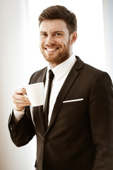 Business concept. Successful young businessman at work. Manager standing in office happy drinking coffee from cup. Man smiling in suit indoors on glass window background