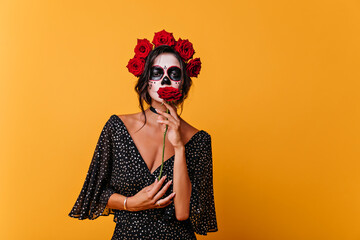 Romantic dead girl in black dress holding red rose. Indoor portrait of female zombie wears flower wreath.