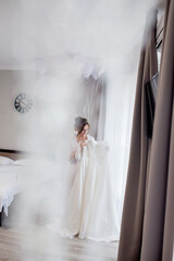 Young woman in silk bathrobe touching her wedding dress and smiling while standing near the window. Wedding day. Beauty, fashion. Happiest bridal day. Enjoy every moment.