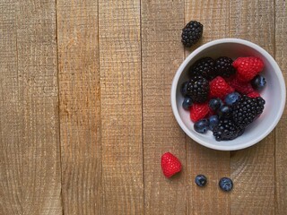 in a soft light table top view, a vertically flat lay a white bowl with colorful berries on brown wooden boards, on the table, in it a fresh mix of blueberries, raspberries and blackberries
