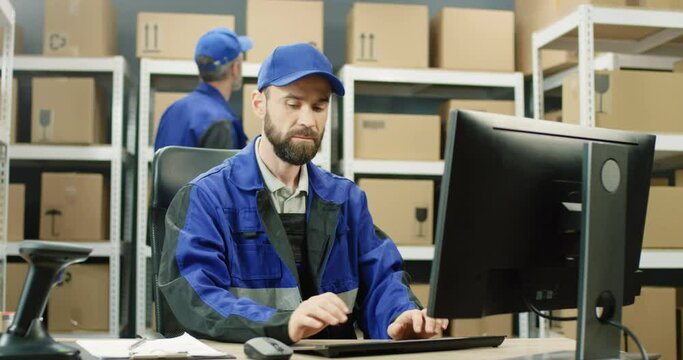 Handsome Caucasian postman in iniform and cap sitting at table in postal office store and working at computer. Male courier typing on keyboard in storage of parcels. Man browsing online.