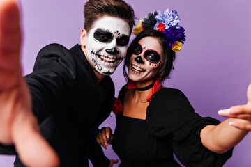 Positive zombie guy making selfie in studio with girlfriend. Happy friends in masquerade costumes...