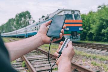 The guy charges a smartphone through a portable charger on the background of the train.