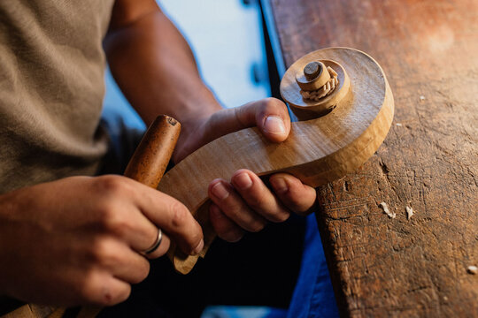 Cropped faceless craftsman carving swirl of violin head while working with wooden detail using bit at table in workshop