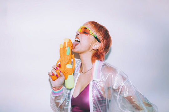 Stylish young female in retro futuristic outfit licking water pistol with closed eyes against gray background