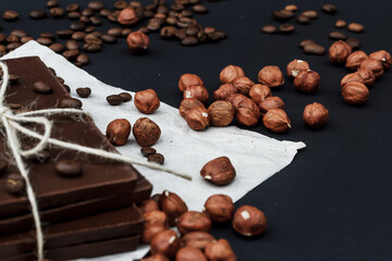 Black chocolate on a black background close-up. Hazelnuts and coffe beans are scattered in the background. Free space in the background. Image of Flat lay. World Chocolate Day.