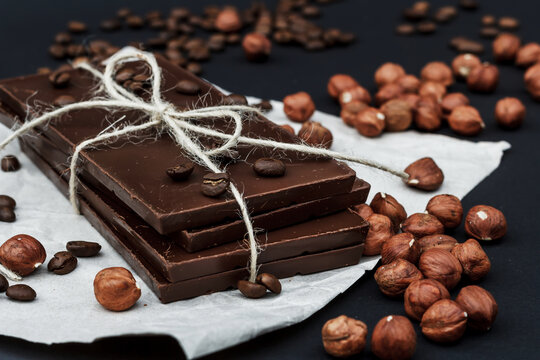 Black chocolate on a black background close-up. Hazelnuts and coffe beans are scattered in the background. Free space in the background. Image of Flat lay. World Chocolate Day.
