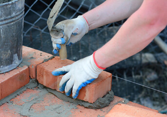 A mason in protective gloves is laying bricks using a trowel, mortar and level line or string to ensure the brickwork is upright and level.
