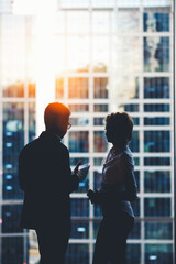 Man and woman professional bankers standing near skyscraper window in modern interior, two business people using digital tablet and mobile phone for teamwork, colleagues discuss ideas after briefing