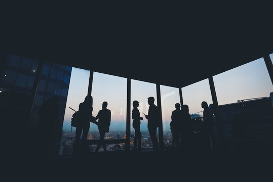 Silhouette Of Group Of Young Skilled Business People Talking Among Themselves While Standing In Modern Office Interior Near Window, Mans And Women's Purposeful Bookkeepers Discuss Ideas After Meeting